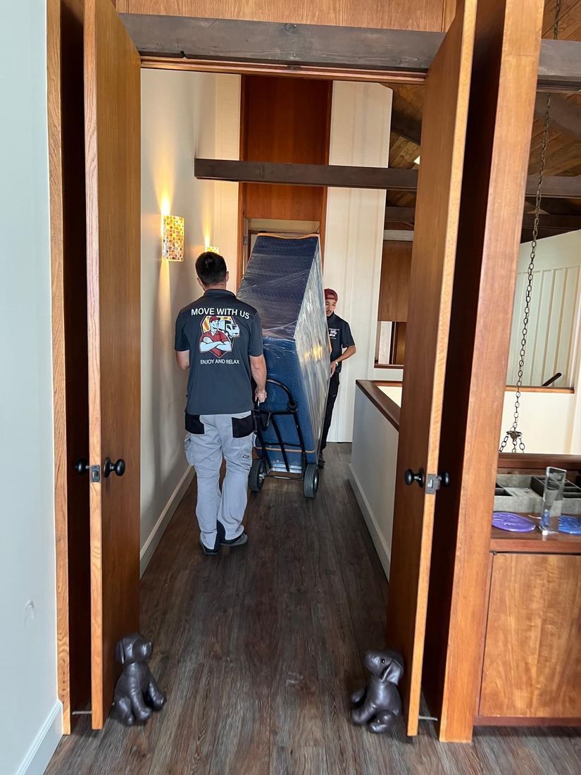 Two movers rolling a wrapped piece of furniture through a narrow hallway
